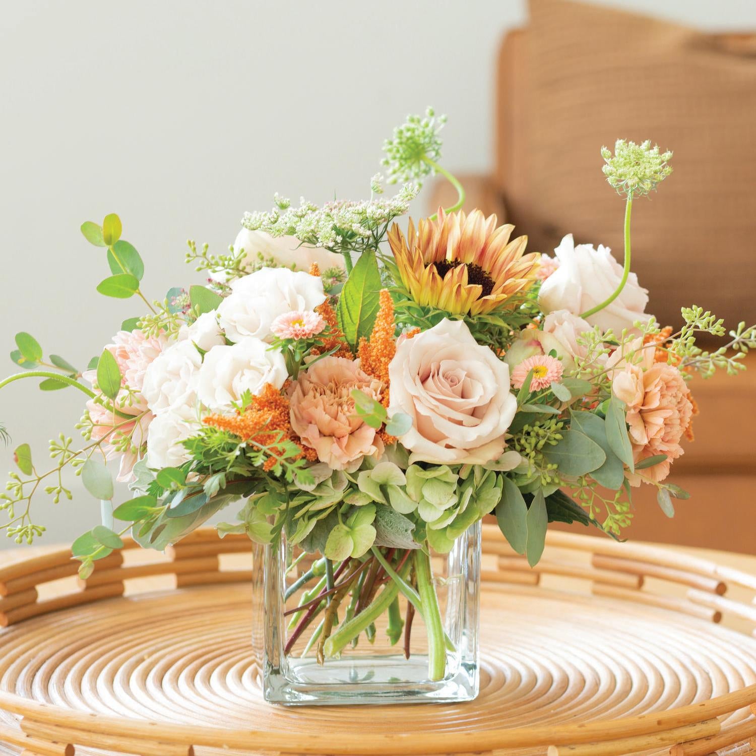 Fresh and elegant floral centerpiece with blush roses, peach carnations, green hydrangeas, and sunlit daisies in a clear glass vase, set upon a natural woven rattan table, creating a warm, inviting home decor atmosphere.
