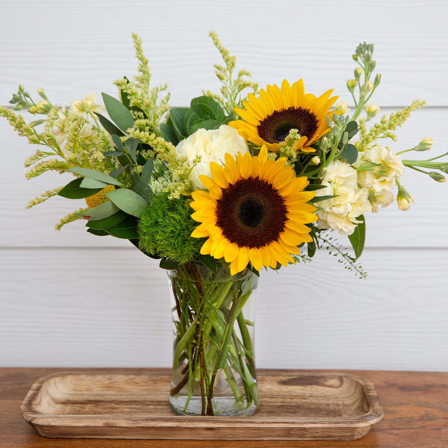 Vibrant bouquet featuring bright sunflowers, white roses, and lush greenery arranged in a clear prism vase, set upon a wooden tray, creating a cheerful display for any interior space, available at 46spruce.com.