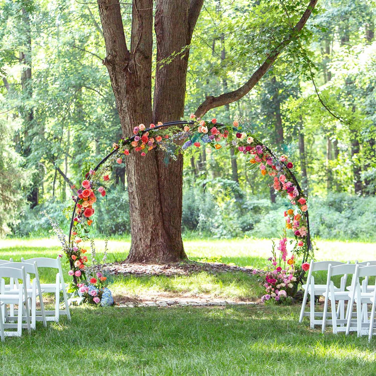 46 & Spruce round outdoor wedding arch: vibrant full-circle floral arrangement with pink, orange, and purple flowers. Set against lush green trees, creating a perfect outdoor setting.