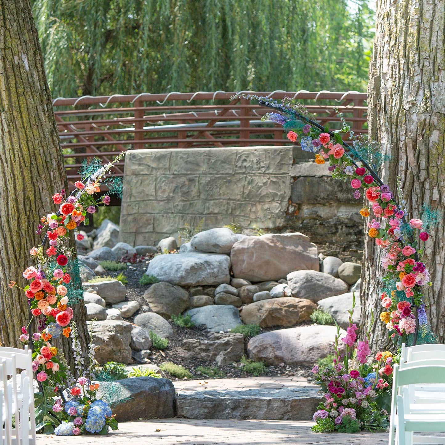 46 & Spruce outdoor wedding arch: vibrant half-circle floral display with pink, orange, and purple flowers. Set against a rocky path and a rustic bridge backdrop.