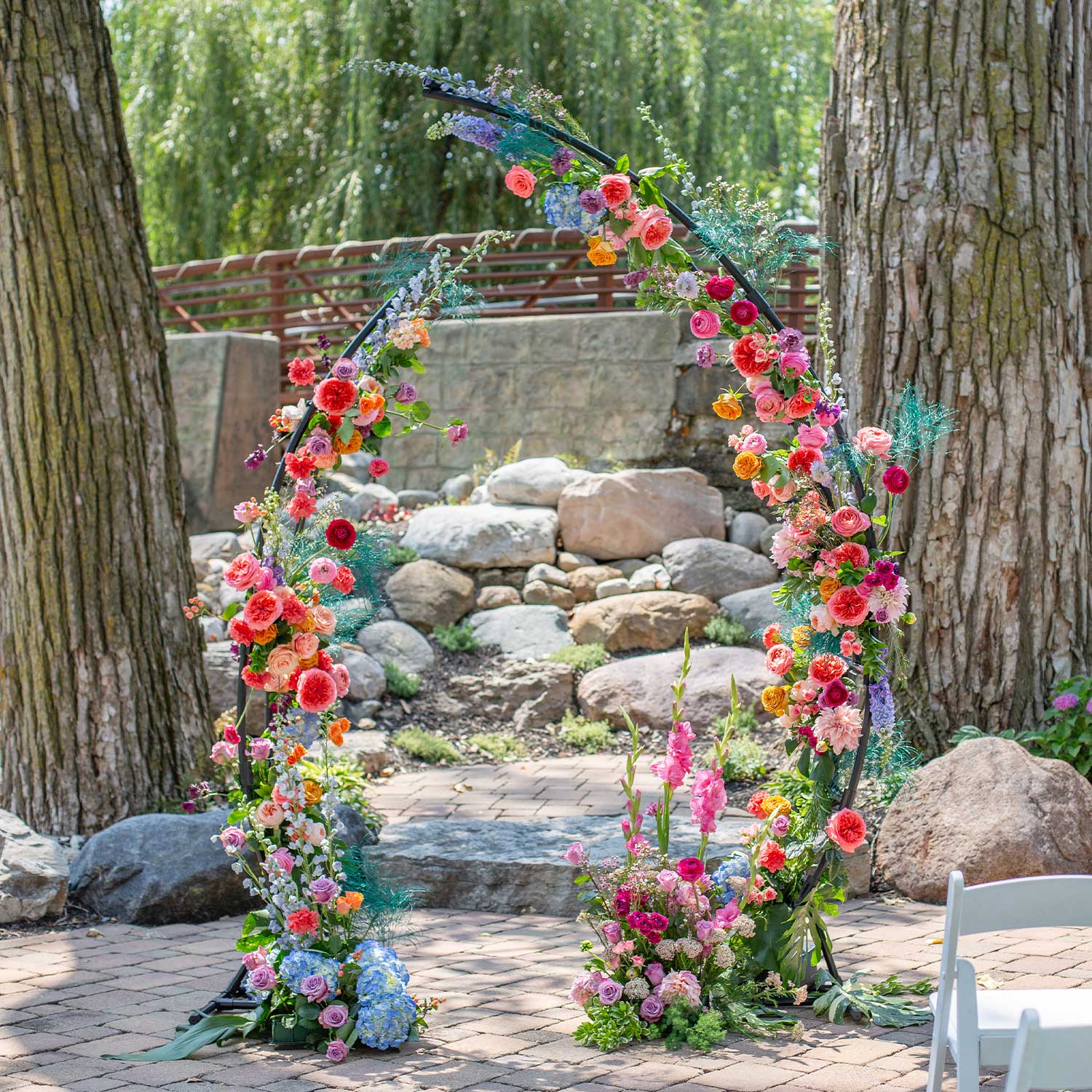 A half round 46 & Spruce floral arch adorned with colorful flowers stands at a pathway entrance, set against a natural backdrop with trees, a fence, and rocks.