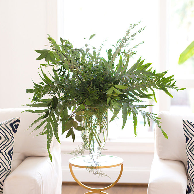 A lush greenery arrangement in a transparent 10 7/8 Mia vase on a gold-rimmed round table between two white chairs with decorative pillows, in a bright room with a window.