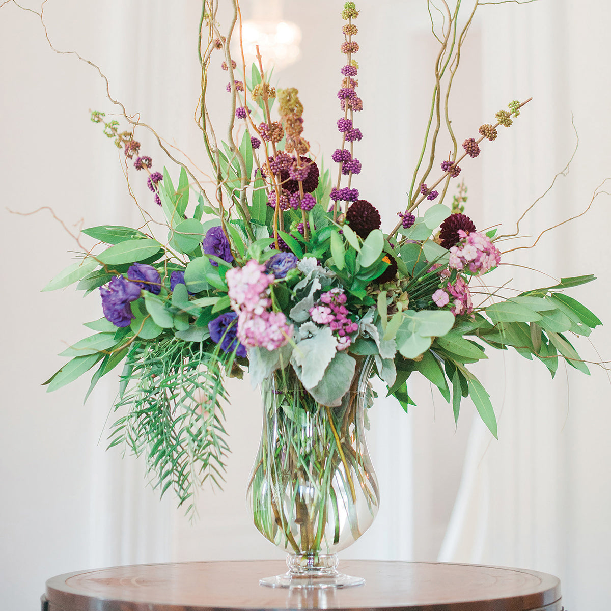 An elaborate bouquet in the 14 inch Charlotte vase, with tall, twisting branches, deep purple flowers, and vibrant greenery, creating a dramatic and elegant centerpiece on a round wooden table, with a soft, neutral backdrop.