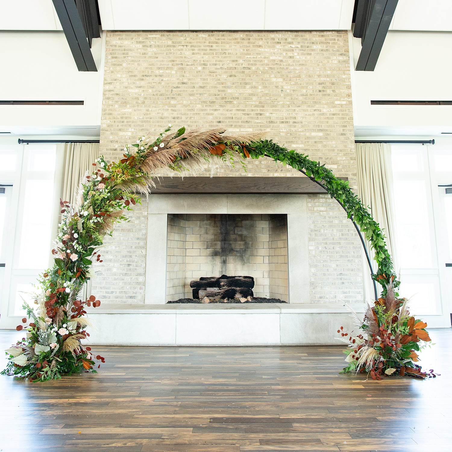 46 & Spruce round wedding arch: elegant half-circle floral design featuring natural greens, brown leaves, and soft flowers. Positioned in front of a fireplace indoors.