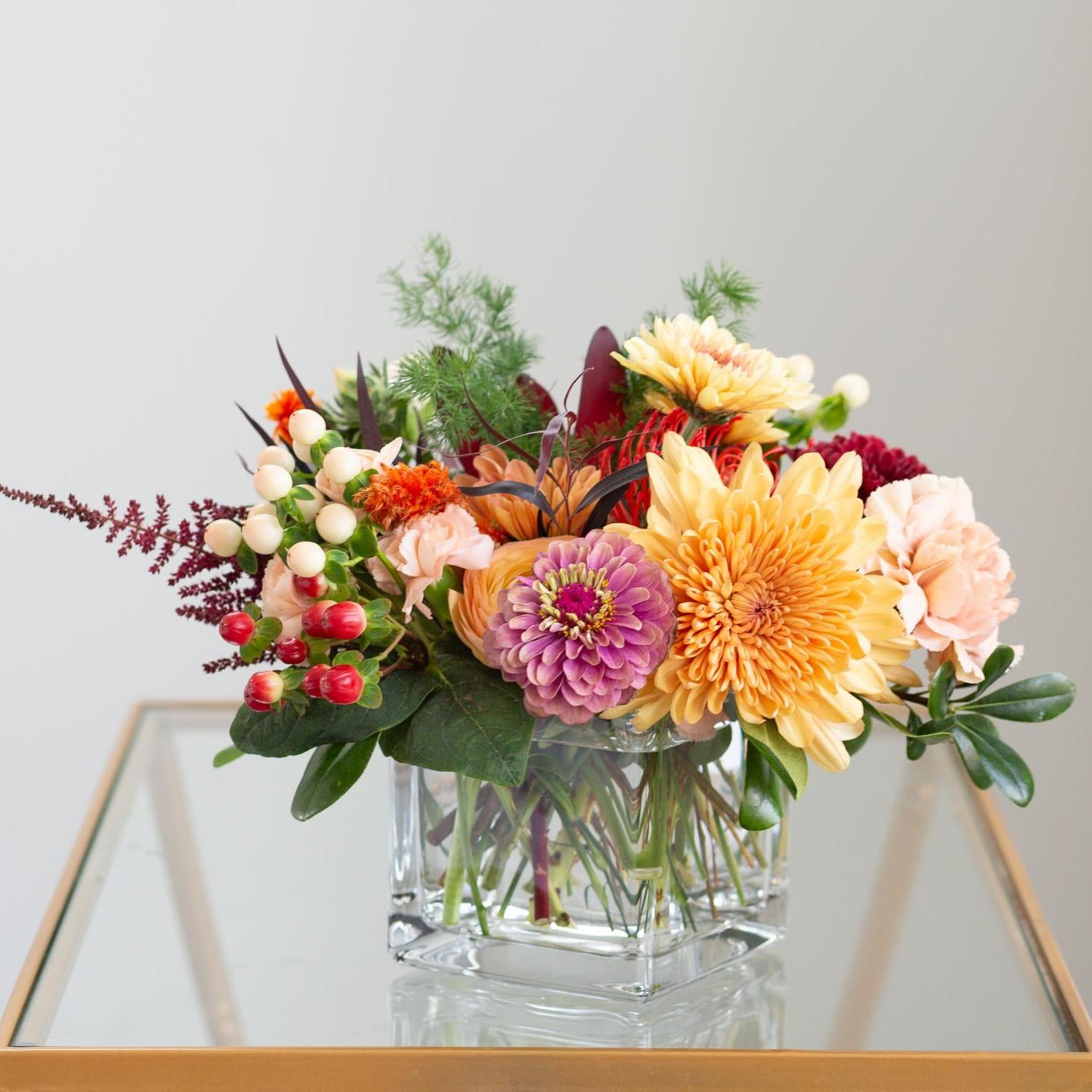 Vibrant autumnal flower arrangement in a square glass vase, featuring purple dahlias, peach carnations, orange mums, and accents of red hypericum berries, on a reflective glass tabletop with a gray wall background.
