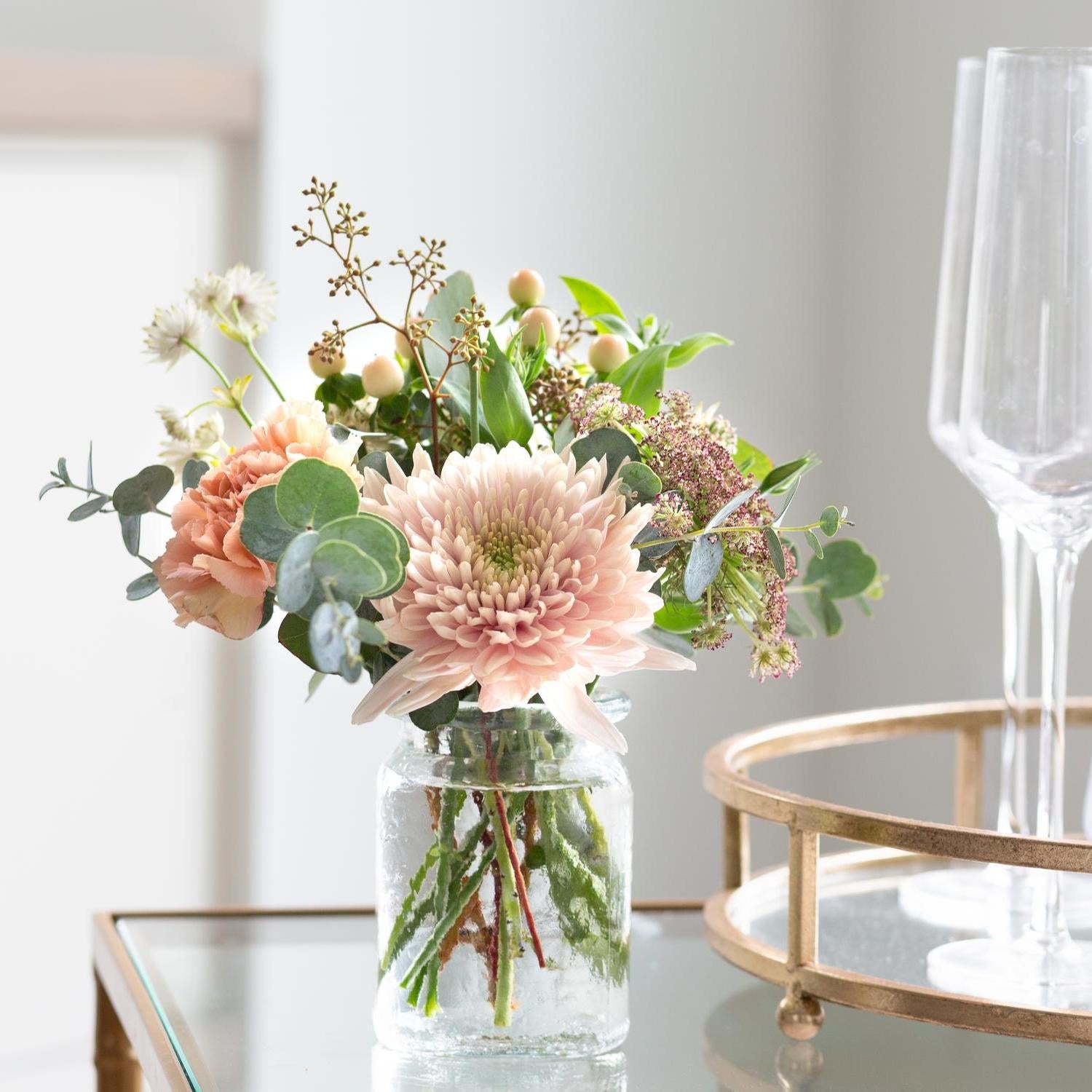 Elegant floral arrangement featuring peach roses, blush chrysanthemums, and assorted greenery in a clear hammered glass jar vase, on a modern glass table beside an empty crystal champagne flute, with a wooden serving tray in soft focus in the background.