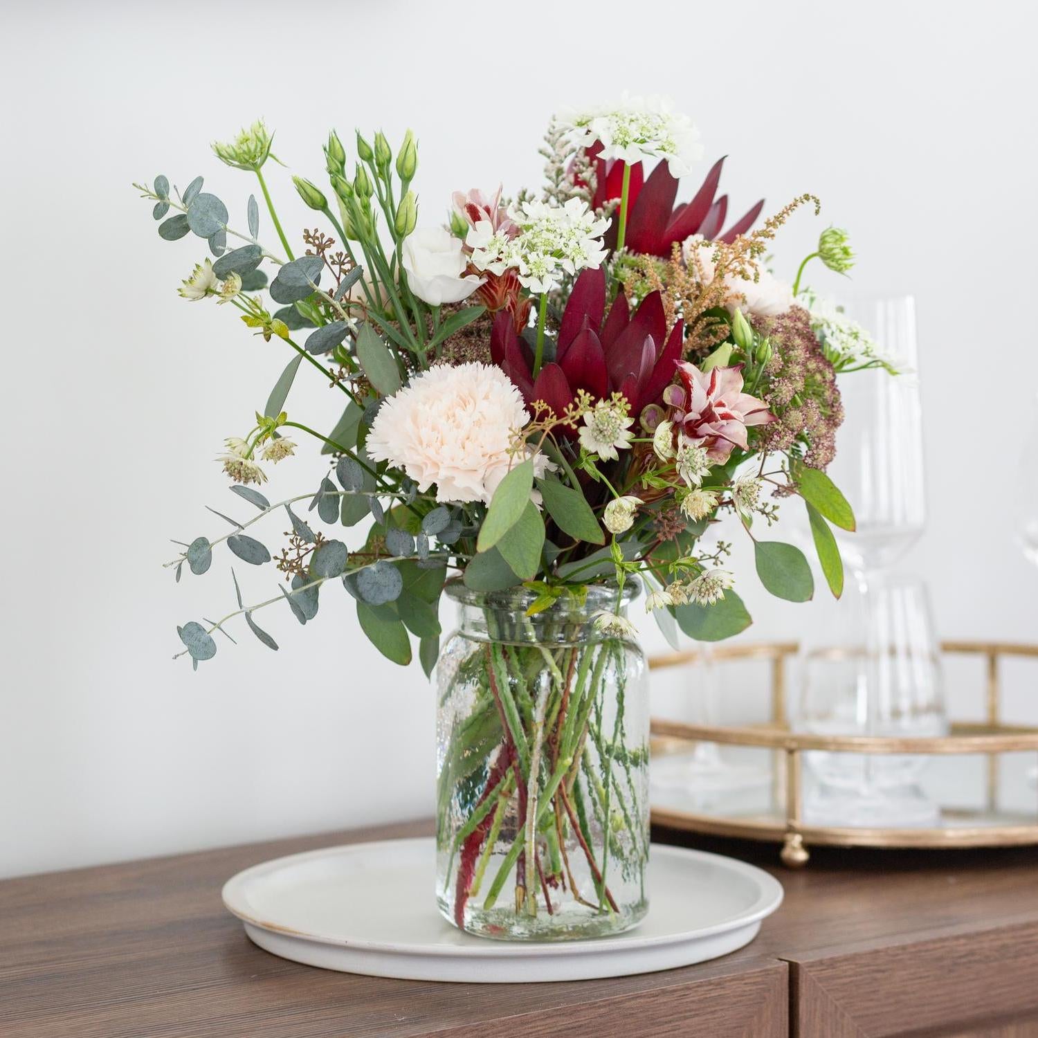 Luxurious bouquet with deep red magnolias, blush carnations, white roses, and lush greenery arranged in a textured glass jar vase, displayed on a wooden sideboard next to white ceramic plates and a reflective gold-rimmed serving tray with glassware in the background.