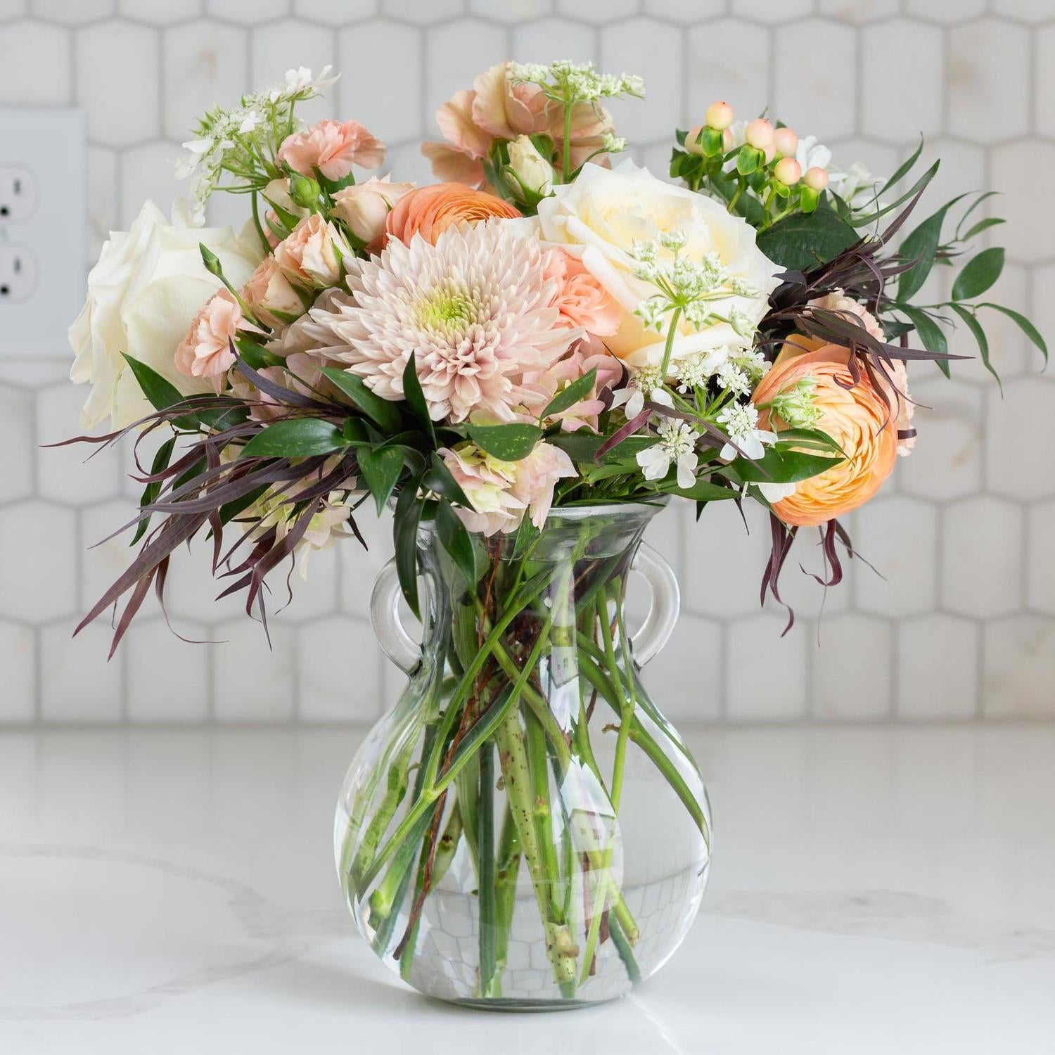 Elegant glass vase filled with blush pink roses and coral blooms against a tiled backsplash.