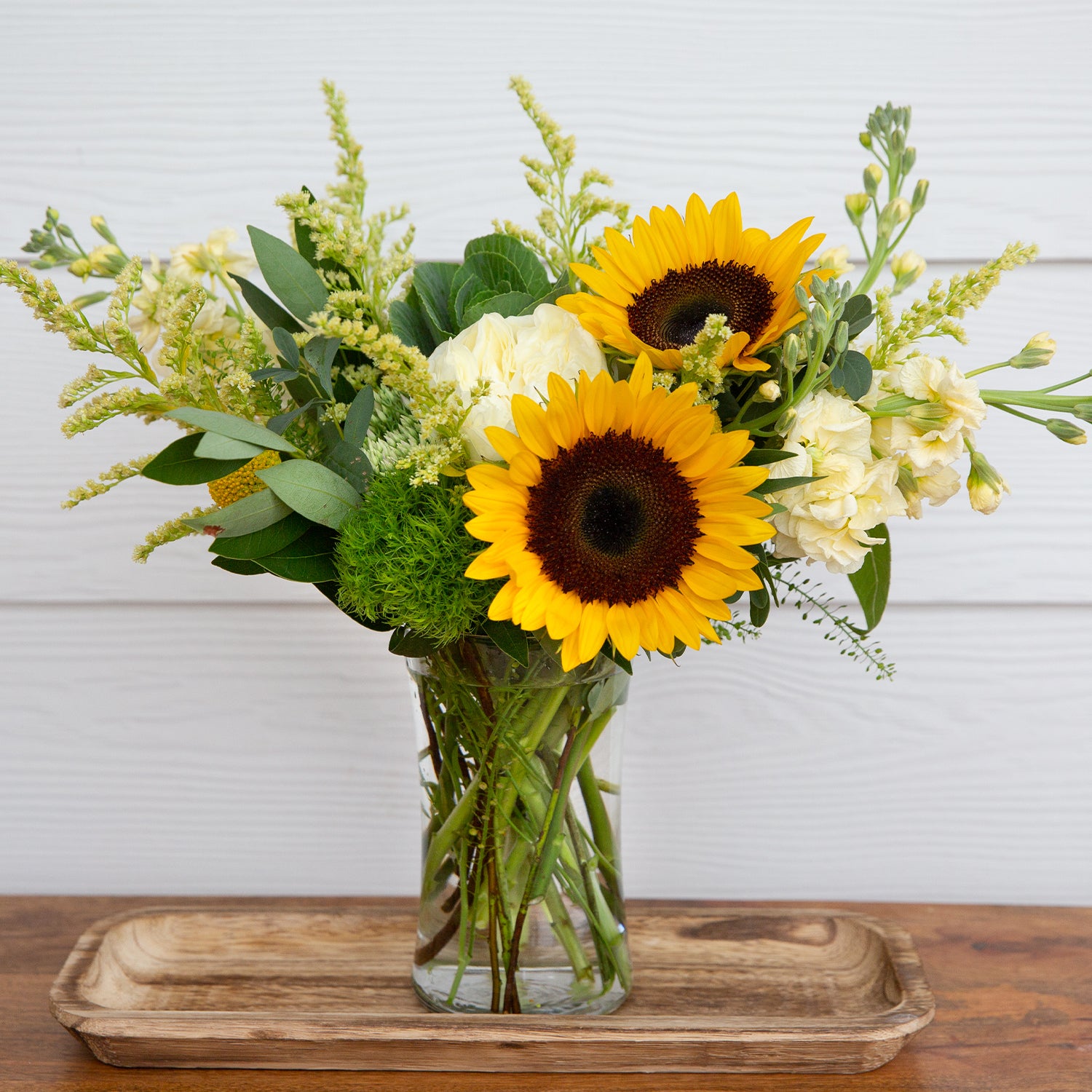 Vibrant bouquet featuring bright sunflowers, white roses, and lush greenery arranged in a clear prism vase, set upon a wooden tray, creating a cheerful display for any interior space, available at 46spruce.com.