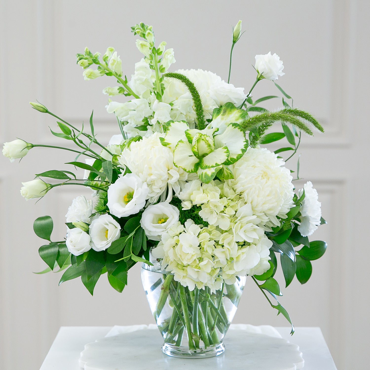 A 5 3/4 inch Serenity vase filled with an all-white floral arrangement, including lush peonies, delicate lisianthus, hydrangeas, and ornamental greenery, displayed on a marble tabletop with a blurred light interior background.