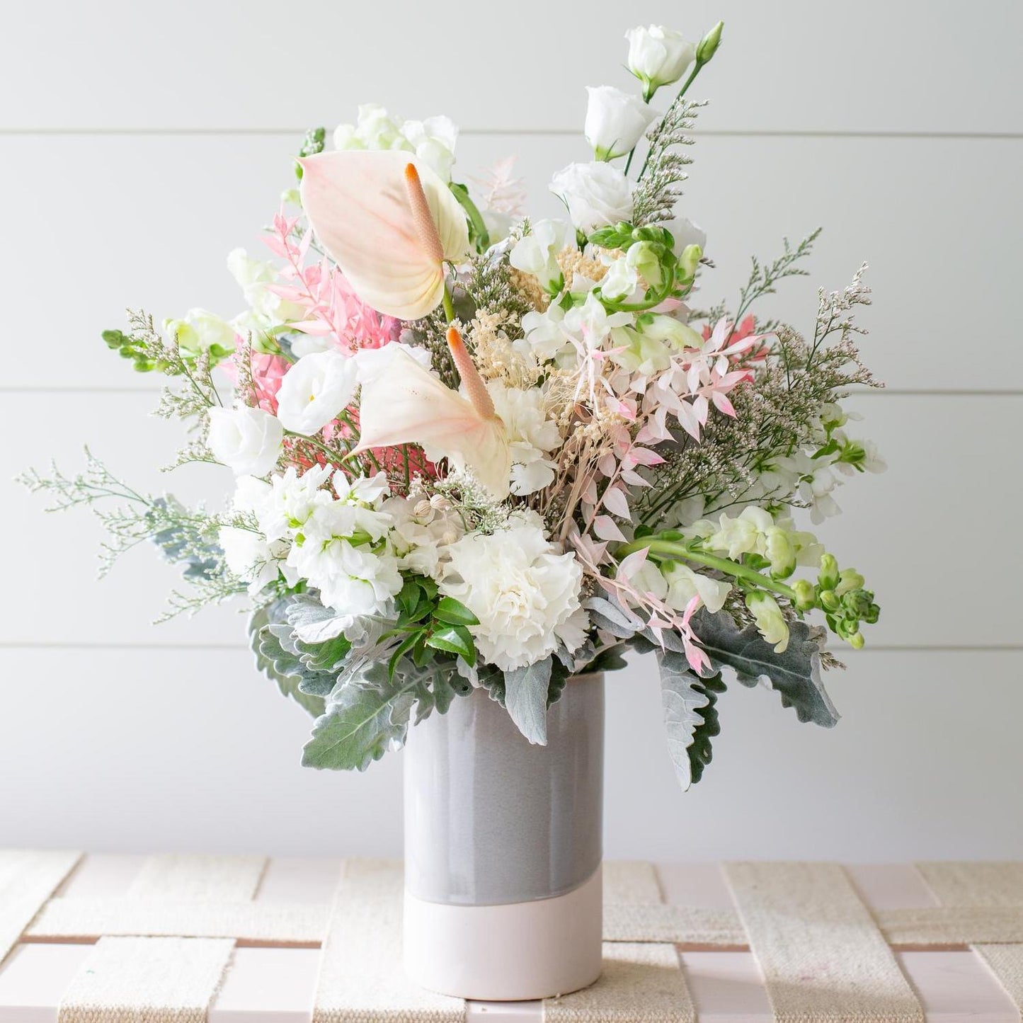 A lush bouquet of flowers, featuring pale pink calla lilies, white roses, and an assortment of greenery and filler flowers, is elegantly arranged in a grey ceramic essential vase placed on a light-colored wooden surface against a white horizontal panel backdrop.