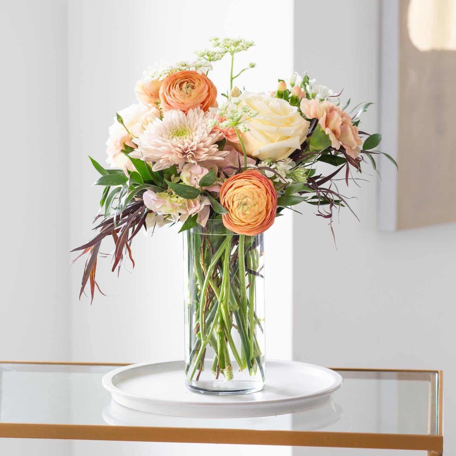 Tall cylinder vase displaying a sophisticated arrangement of peach ranunculus, blush dahlias, and delicate white accents in a modern living space.