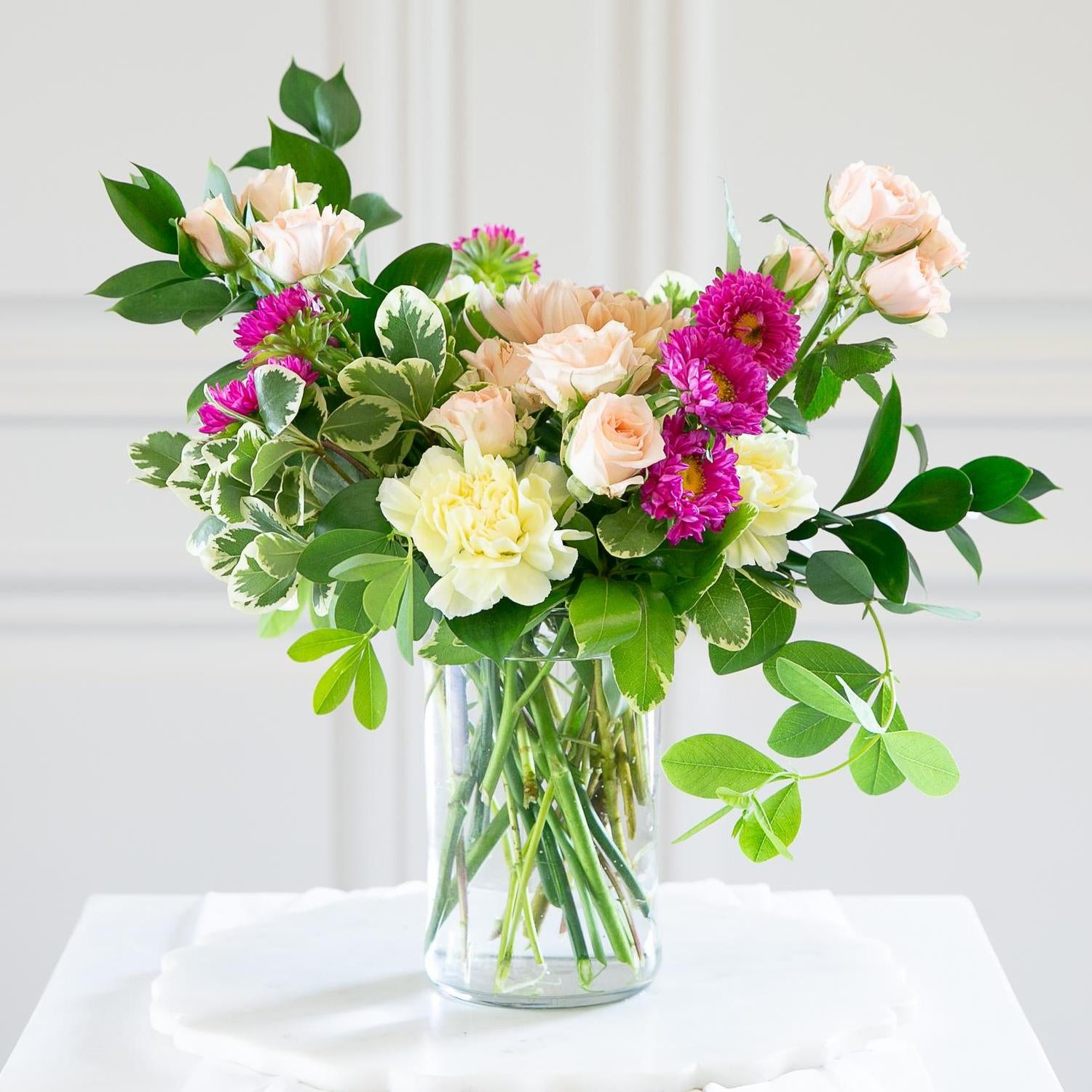 Glass cylinder vase with a vibrant selection of pink roses, purple asters, and lush greenery on a marble table.
