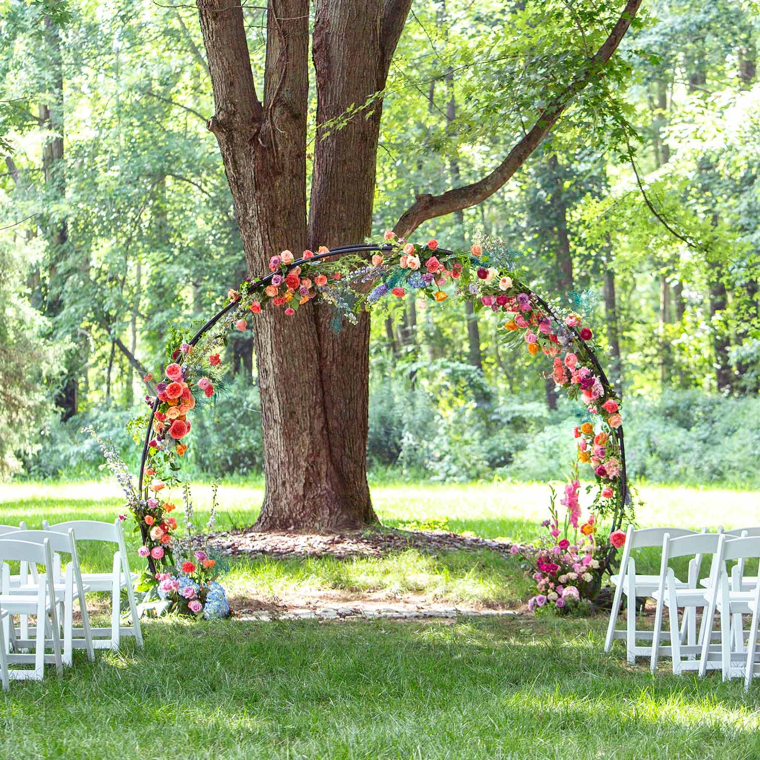 46 & Spruce round outdoor wedding arch: vibrant full-circle floral arrangement with pink, orange, and purple flowers. Set against lush green trees, creating a perfect outdoor setting.