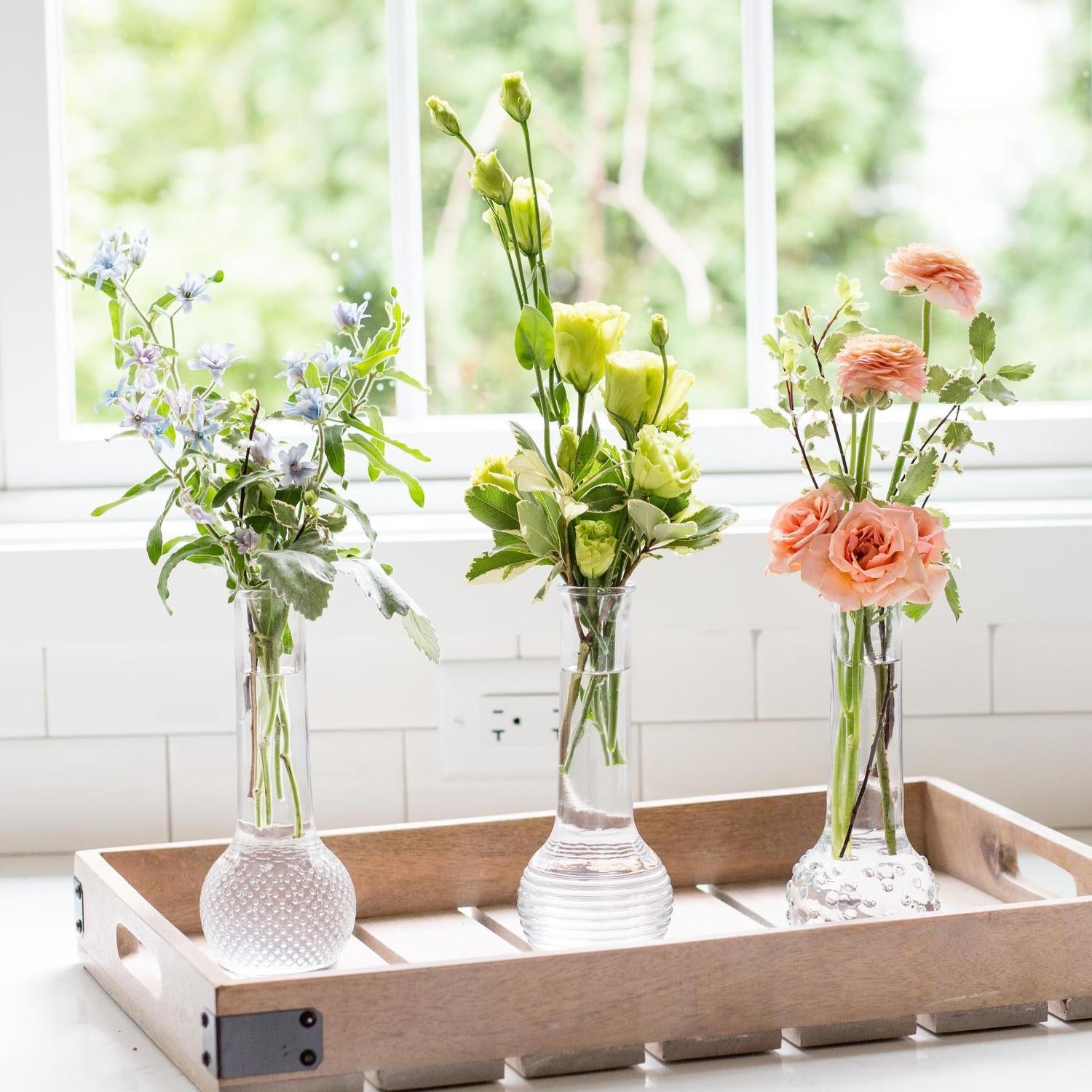 The bud vase assortments are filled with a variety of flowers including blue forget-me-nots, green lisianthus, and peach roses, placed on a wooden tray on a white windowsill, offering a view of green foliage outside.