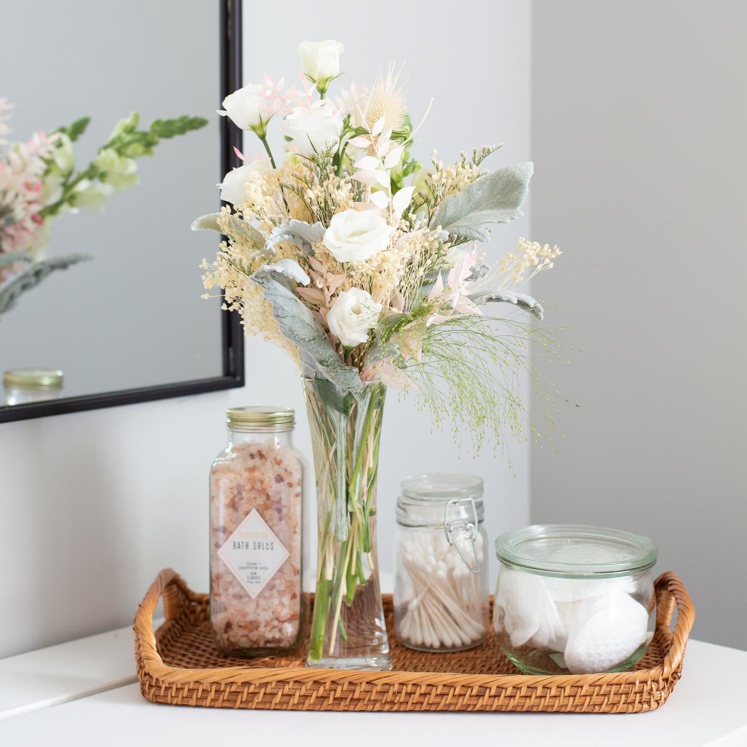 An arrangement within the vase featuring white roses, pale pink flowers, and assorted greenery, placed on a wicker tray beside bath salts and cotton balls, with a bathroom mirror partially visible in the background.