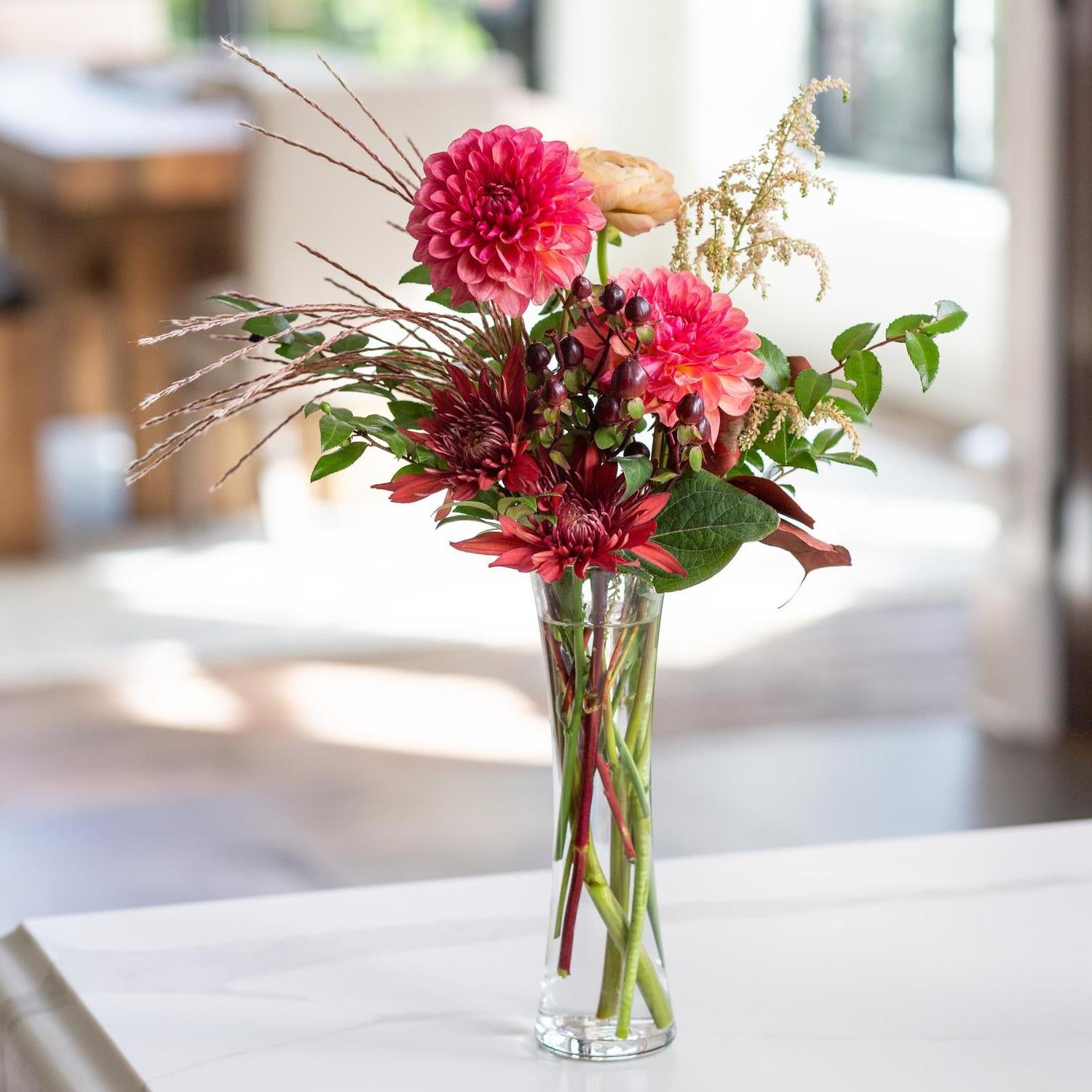 A Selena Glass Bud Vase filled with a vibrant arrangement of red and pink dahlias, complemented by dark red berries, green leaves, and decorative grasses, sitting on a white surface with a blurred indoor background.