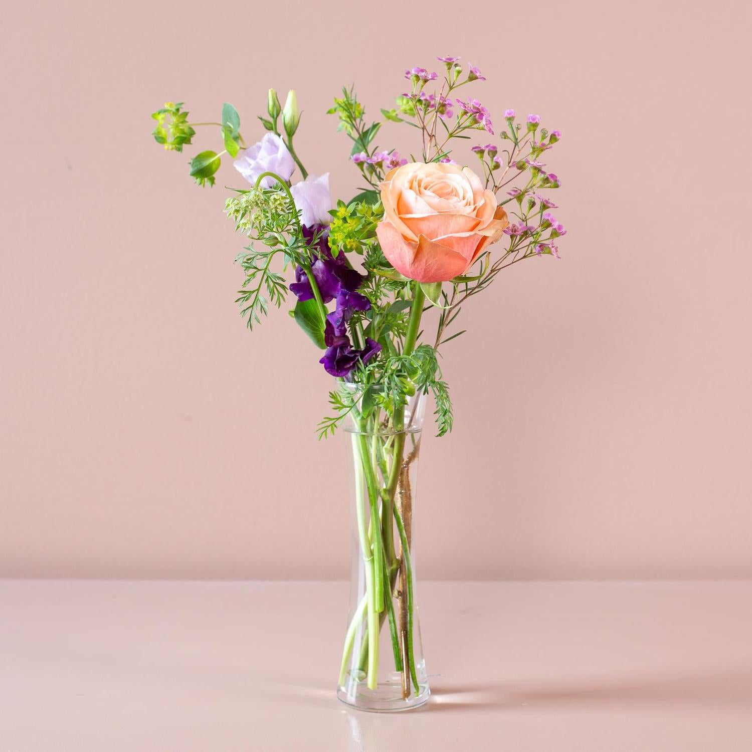 The vase holds a delicate display of flowers, including a peach-colored rose, purple flowers, and greenery, set against a pale pink background.