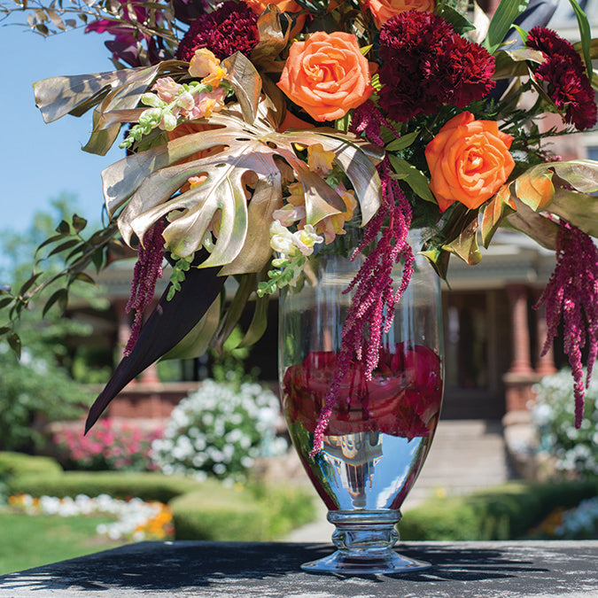 A colorful floral arrangement in a clear Mia Vase with orange roses, purple carnations, greenery, and submerged maroon petals, set against a garden backdrop.