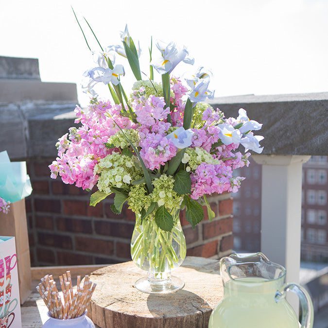 A colorful floral arrangement in the vase, with pink hydrangeas, light blue irises, and green foliage, displayed outdoors on a wooden surface, with a pitcher of lemonade and city buildings in the background.