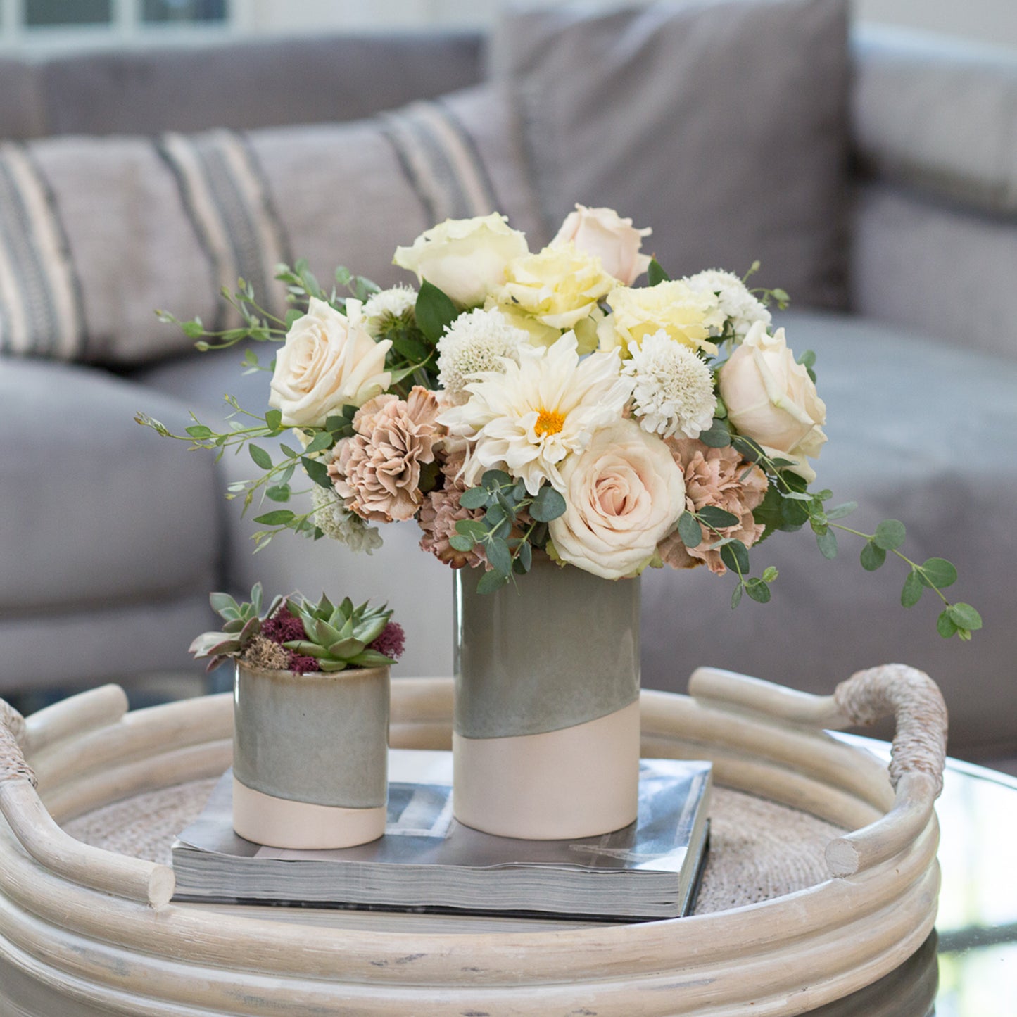 An elegant floral arrangement in a 6.5-inch ceramic essential vase accompanied by a smaller 4-inch version, both in a dual-tone design with pale green and blush pink. The larger vase holds a bouquet of creamy roses, pale yellow flowers, and soft greenery, while the smaller one contains a succulent with accents of deep purple. This display sits atop a round wicker tray on a glass table, with a cozy grey sofa adorned with pillows in the background.